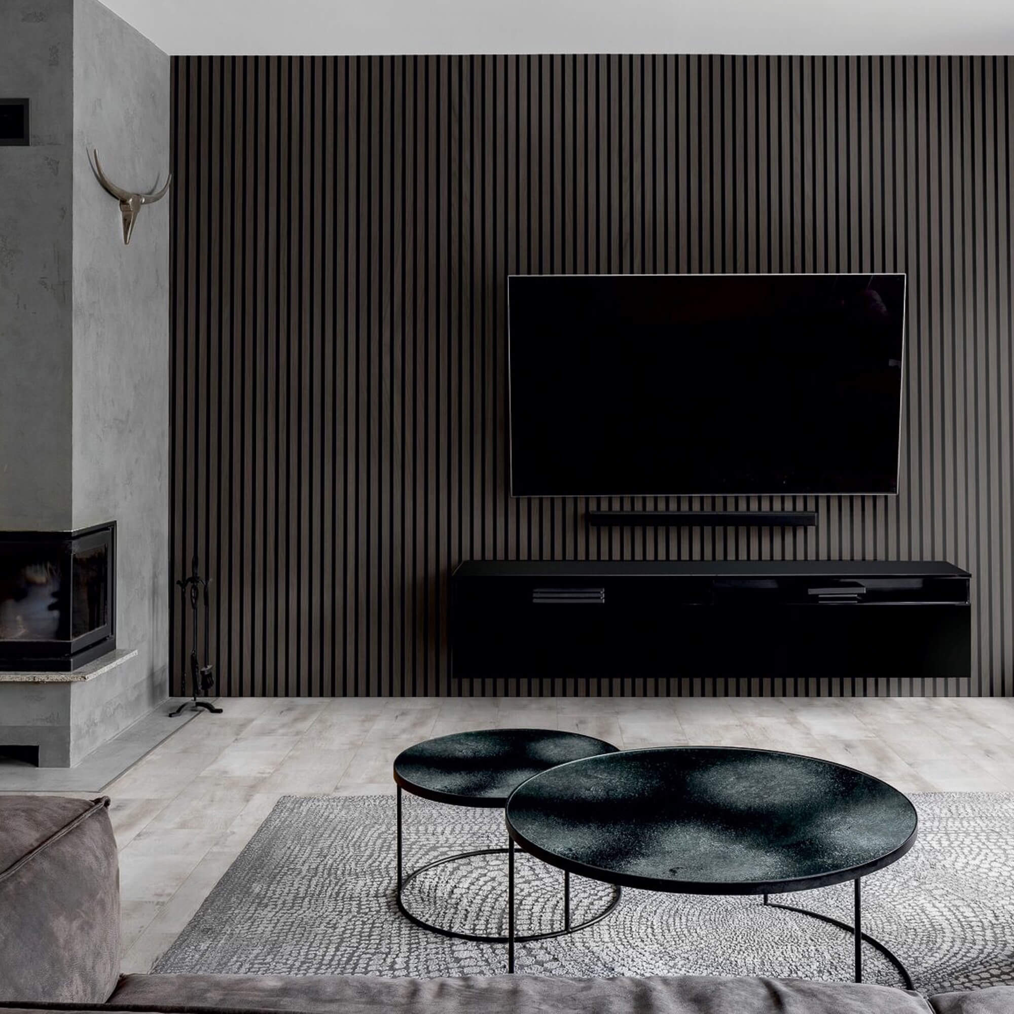Der große Flachbildfernseher hängt mittig an einer dunklen, vertikal-geriffelten Wand. Davor zwei runde niedrige Tische auf hellem Teppich; links ein Kamin und ein schwebendes schwarzes Sideboard in minimalistischem Wohnzimmer.