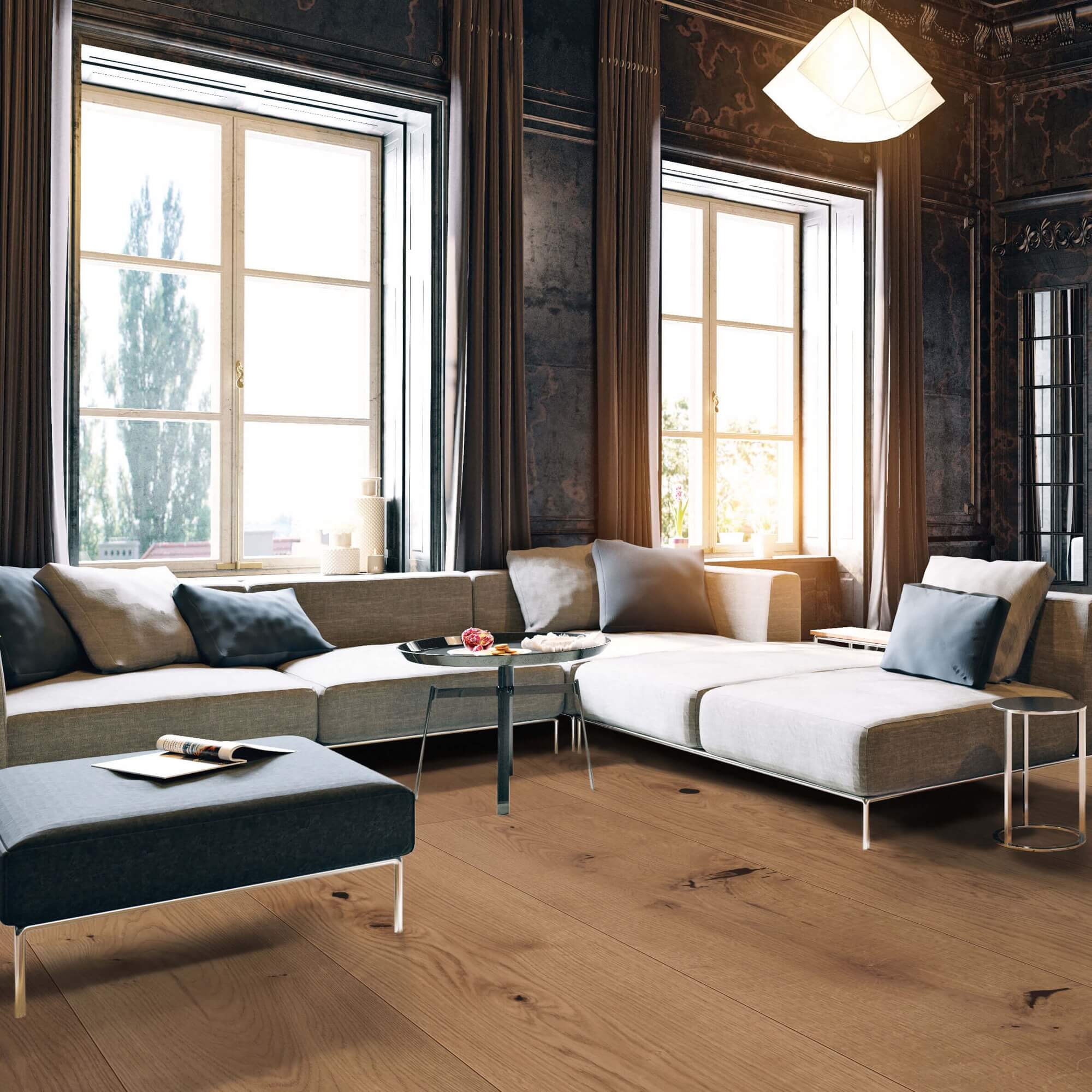 Großes hellgraues Ecksofa steht im Vordergrund; Kissen liegen darauf; Sonnenlicht fällt durch zwei hohe Fenster; dunkle Vorhänge, hölzerner Parkettboden und ein kleiner Couchtisch im Raum.