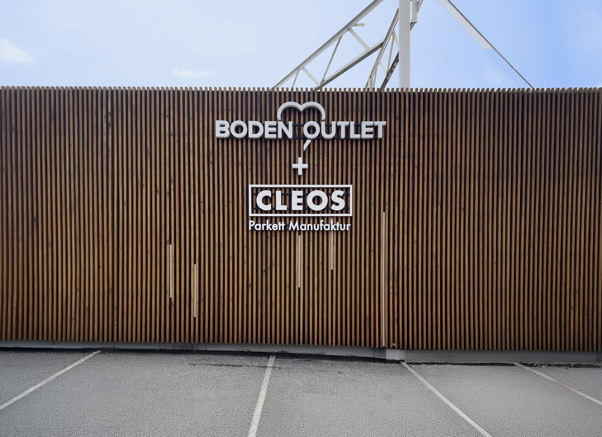 Eine orangefarbene Ladenfront mit großem Schriftzug "BODEN OUTLET", die Eingangsfront präsentiert Sortiment-Labels; ruhiger Parkplatz davor und blauer Himmel im Hintergrund.