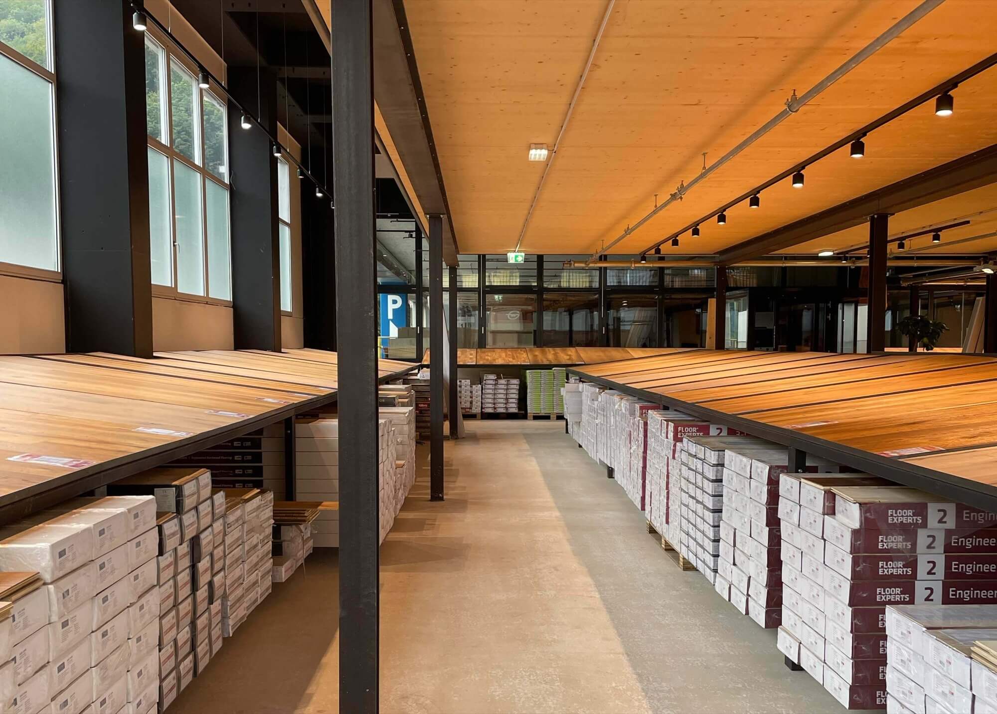 Große Holzpaneele liegen schräg auf Ausstellungstischen; darunter gestapelte Kartons mit Bodenbelägen; die Szene befindet sich in einem hellen, hohen Lager-Showroom mit Fensterfront.