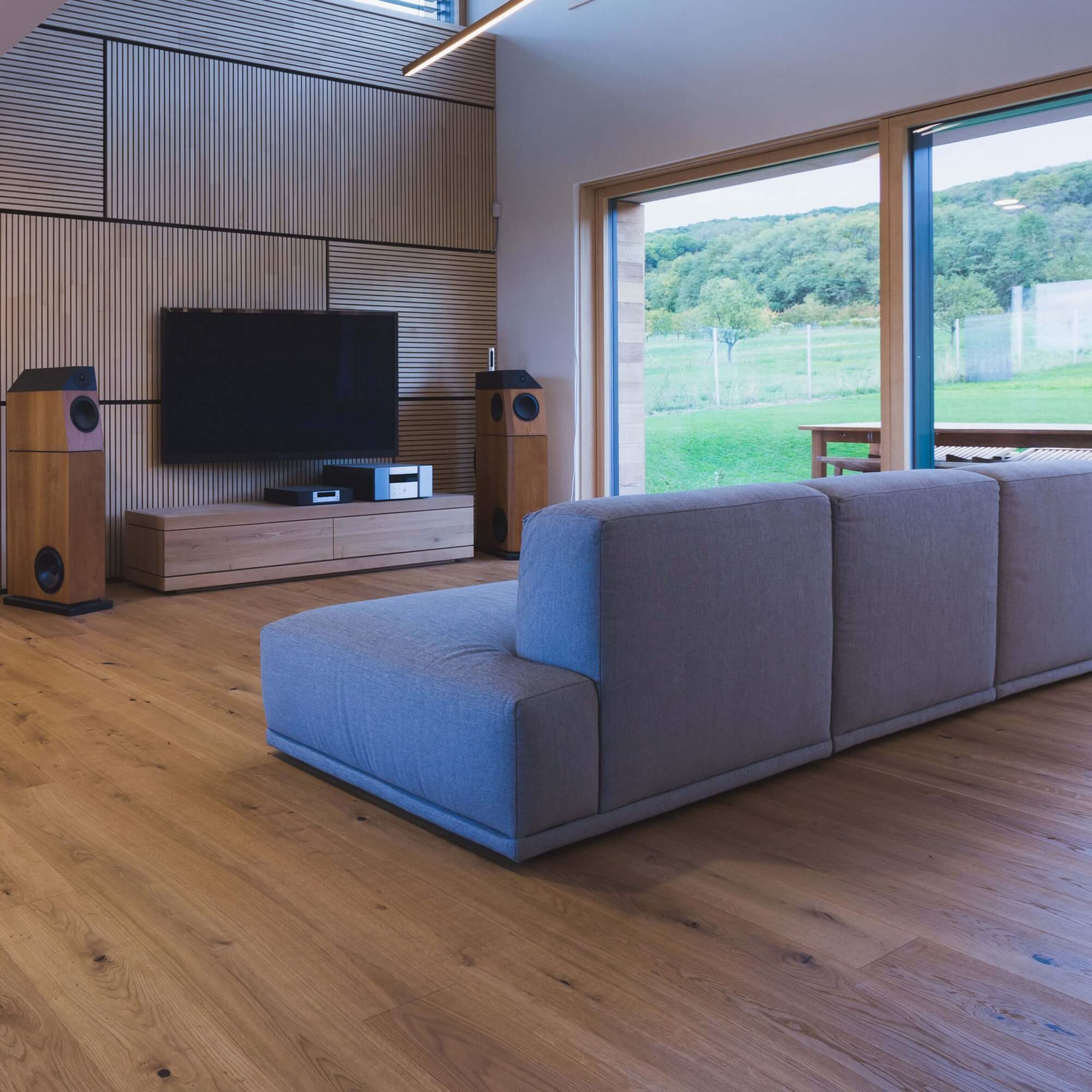 Graue Eckcouch sitzt mittig im Raum, ausgerichtet auf einen an der Holzlattenwand montierten Flachbildschirm; umgeben von Holzboden, Standlautsprechern und großen Glasschiebetüren mit Blick auf grüne Hügel.