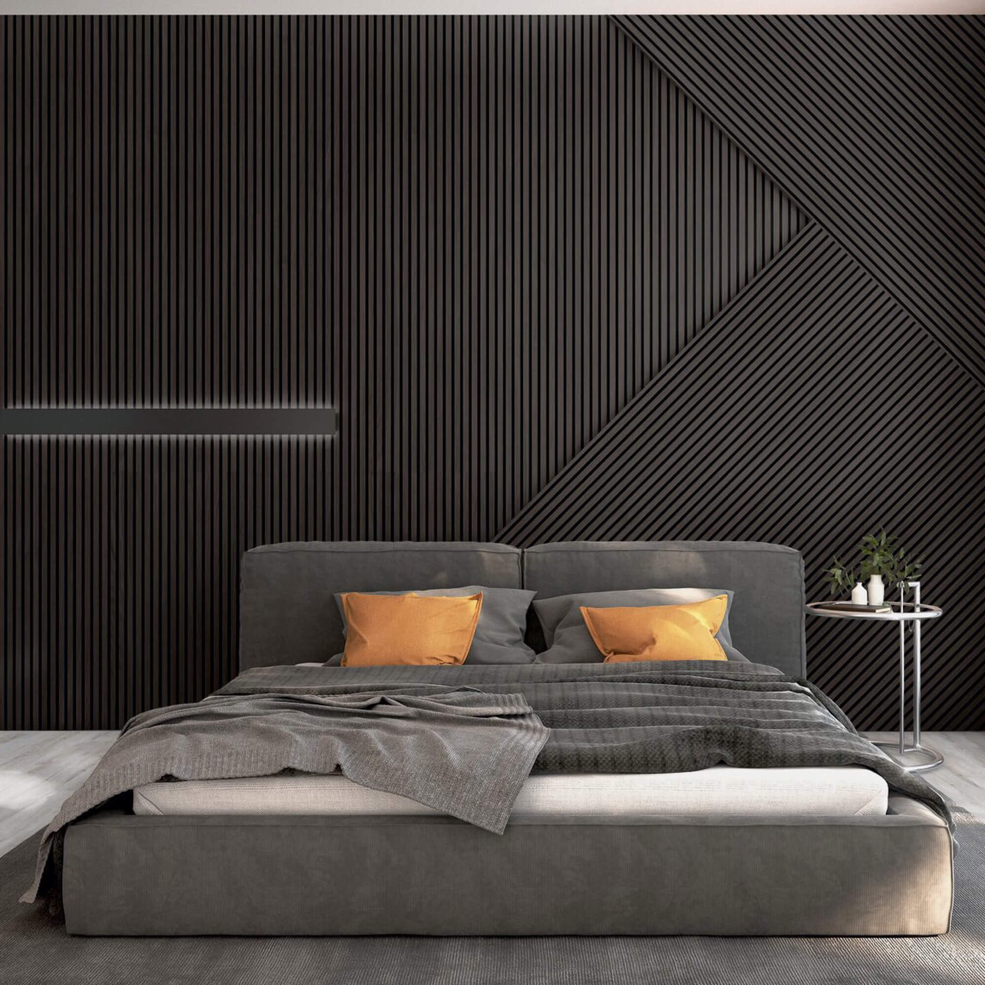 Bett dominiert den Vordergrund, mit grauem Polster, zerknitterter Decke und zwei orangen Kissen; dahinter dunkel strukturierte Wand mit vertikalen und diagonalen Lamellen; rechter Beistelltisch mit Pflanzen.