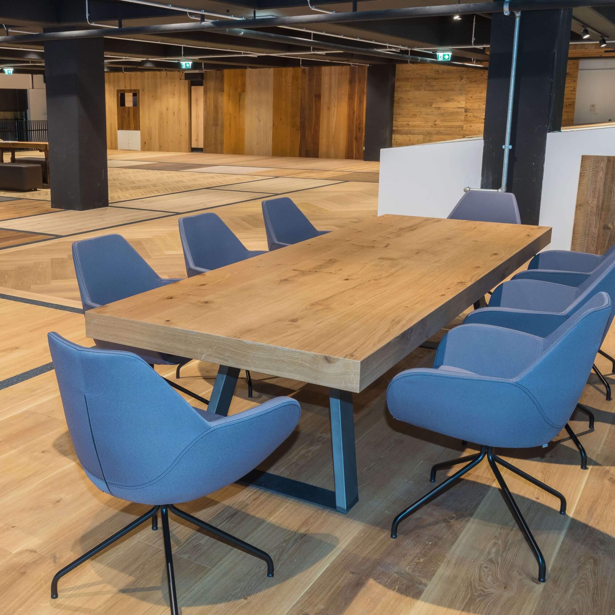Großer massiver Holztisch steht zentral, umgeben von sechs blauen Polsterstühlen; moderne, offene Innenraumgestaltung mit Holzfußboden, dunklen Säulen und dezenter Deckenbeleuchtung.